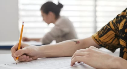 Asmuo sėdi prie stalo ir pieštuku pildo testą; fone matomas kitas žmogus,