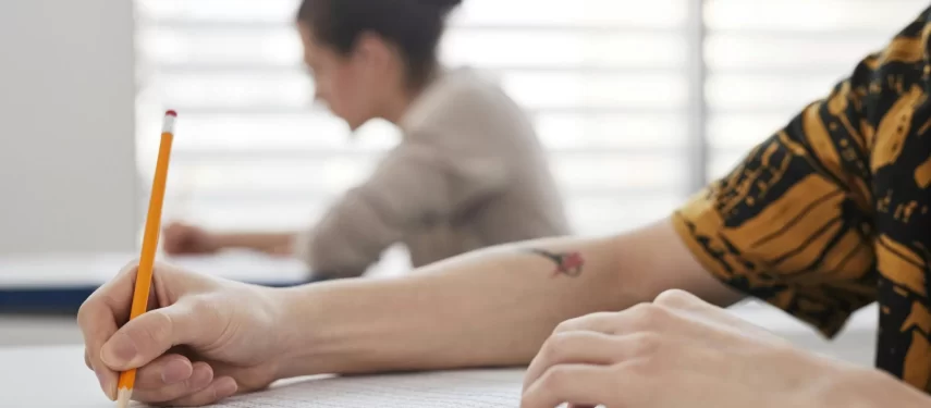 Asmuo sėdi prie stalo ir pieštuku pildo testą; fone matomas kitas žmogus,