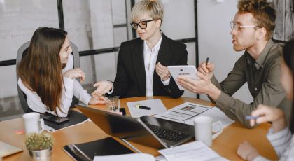 Keturi darbuotojai sėdi prie stalo ir diskutuoja, o ant stalo galima matyti kompiuterį ir daug dokumentų, pexels.com nuotr.