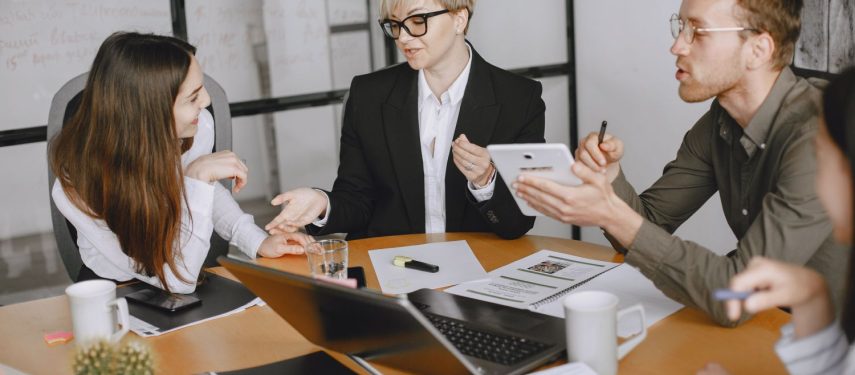 Keturi darbuotojai sėdi prie stalo ir diskutuoja, o ant stalo galima matyti kompiuterį ir daug dokumentų, pexels.com nuotr.
