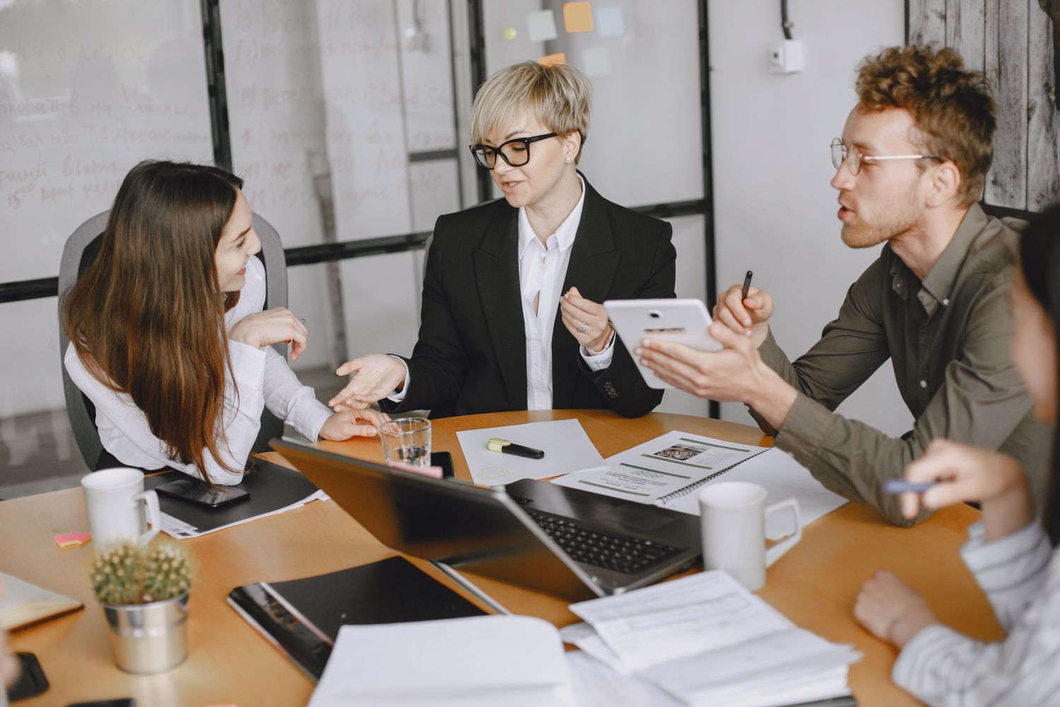 Keturi darbuotojai sėdi prie stalo ir diskutuoja, o ant stalo galima matyti kompiuterį ir daug dokumentų, pexels.com nuotr.