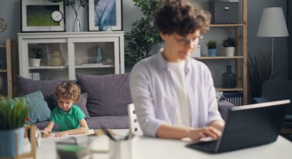 Moteris sėdi prie stalo ir naudojasi kompiuteriu, už jos vaikas sėdi šalia sofos ir piešia, pexels.com nuotr.