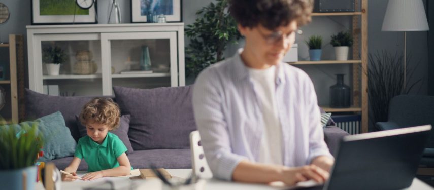Moteris sėdi prie stalo ir naudojasi kompiuteriu, už jos vaikas sėdi šalia sofos ir piešia, pexels.com nuotr.