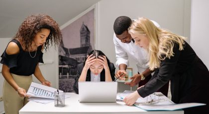 Prie stalo, ant kurio padėtas nešiojamasis kompiuteris, sėdi moteris susiėmusi galvą rankomis, aplink ją susibūrę trys bendradarbiai su dokumentais, pexels.com nuotr.