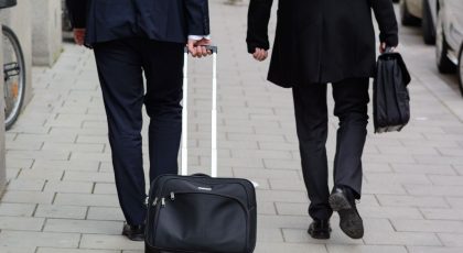 Galima matyti dviejų žmonių kojas, abu dėvi juodas kostiumines kelnes. Vienas iš jų rankoje neša portfelį, o kitas - traukia lagaminą, pexels.com nuotr.