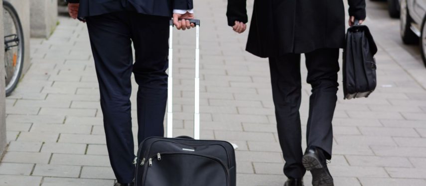 Galima matyti dviejų žmonių kojas, abu dėvi juodas kostiumines kelnes. Vienas iš jų rankoje neša portfelį, o kitas - traukia lagaminą, pexels.com nuotr.