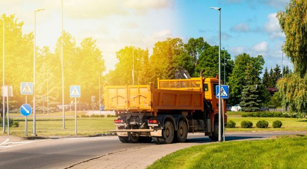 Saulėtą dieną miesto gatve važiuoja oranžinis savivartis, aplink galima matyti pėsčiųjų perėjų ženklus, gatvės žibintus, medžius ir žalią veją, asociatyvi Canva nuotr.