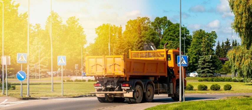 Saulėtą dieną miesto gatve važiuoja oranžinis savivartis, aplink galima matyti pėsčiųjų perėjų ženklus, gatvės žibintus, medžius ir žalią veją, asociatyvi Canva nuotr.