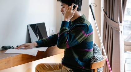 Regos negalią turintis vyras su ausinėmis sėdi prie stalo ant kurio padėti lapai ir kompiuteris, o šalia stalo į sieną atremta baltoji lazdelė, pexels.com nuotr.