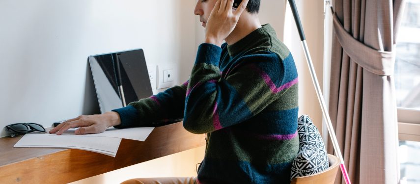Regos negalią turintis vyras su ausinėmis sėdi prie stalo ant kurio padėti lapai ir kompiuteris, o šalia stalo į sieną atremta baltoji lazdelė, pexels.com nuotr.