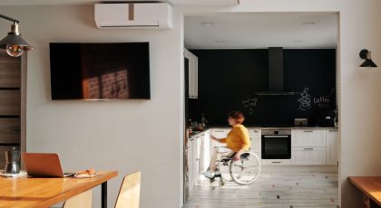 Šviesiame, moderniai įrengtame būste neįgaliojo vežimėliu judantis žmogus yra virtuvėje, nuotraukoje dar galima matyti stalą, kėdes, nešiojamąjį kompiuterį ir televizorių, pexels.com nuotr.