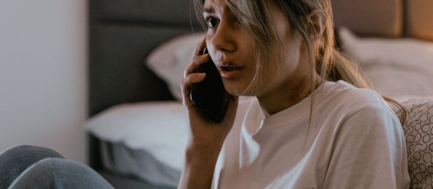 Susirūpinusi moteris sėdi prie lovos ir kalba mobiliuoju telefonu, pexels.com nuotr.