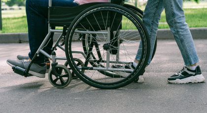 Nuotraukoje matoma tik apatinė žmonių kūno dalis: lauke neįgaliojo vežimėlyje sėdintis žmogus ir šalia jo stovintis kitas asmuo, pexels.com nuotr.