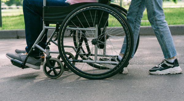 Nuotraukoje matoma tik apatinė žmonių kūno dalis: lauke neįgaliojo vežimėlyje sėdintis žmogus ir šalia jo stovintis kitas asmuo, pexels.com nuotr.