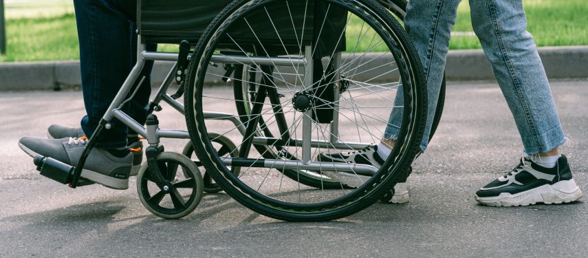 Nuotraukoje matoma tik apatinė žmonių kūno dalis: lauke neįgaliojo vežimėlyje sėdintis žmogus ir šalia jo stovintis kitas asmuo, pexels.com nuotr.