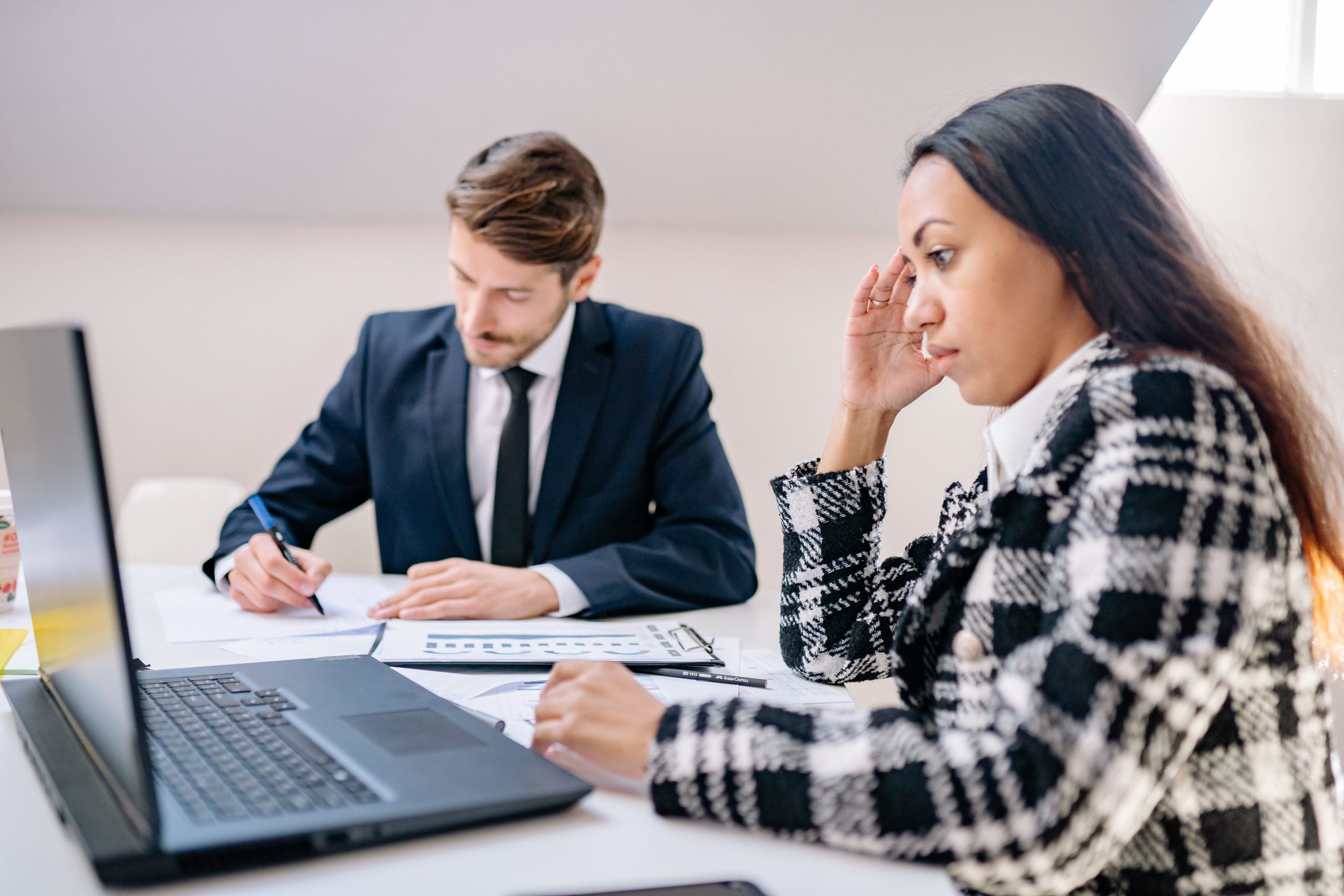 Du darbuotojai sėdi prie stalo, vyras pasirašo dokumentą, o moteris žiūri į kompiuterį, pexels.com nuotr.