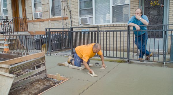 Darbininkas oranžiniais marškinėliais klūpi ir glaisto betoninį šaligatvį, o fone prie tvoros rūkydamas jį stebi vyras mėlynais marškiniais. Betono istorija, filmo stop kadras.