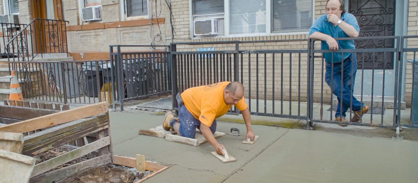 Darbininkas oranžiniais marškinėliais klūpi ir glaisto betoninį šaligatvį, o fone prie tvoros rūkydamas jį stebi vyras mėlynais marškiniais. Betono istorija, filmo stop kadras.