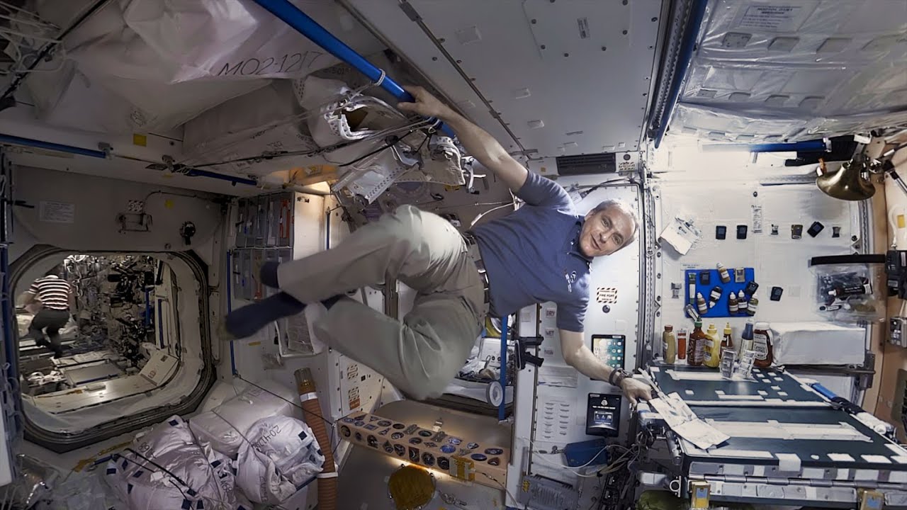 Astronautas mėlynais marškinėliais sklando kosminės stoties viduje, apsuptas įrangos.