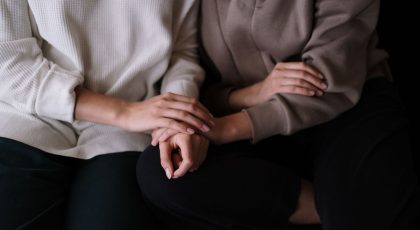 Galima matyti du žmones sėdinčius susiglaudus, ju veidų nematome. Vienas dėvi baltos spalvos megztinį, kitas - rudos. Vieno žmogaus ranka uždėta ant kitos žmogaus rankos, pexels.com nuotr.