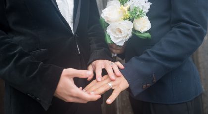 Galima matyti dviejų vyrų viršutinį kūno dalį nuo kaklo iki liemens, abu dėvi kostiumus, vienas laiko gėlių puokštę, o ant kitos rankos piršto galima matyti vedybų žiedą, kito vyro rankos, ant vienos iš jų irgi vestuvinis žiedas, laiko tą ranką ant kurios yra vedybų žiedas, pexels.com nuotr.
