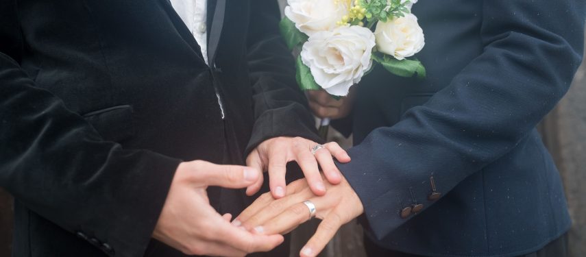 Galima matyti dviejų vyrų viršutinį kūno dalį nuo kaklo iki liemens, abu dėvi kostiumus, vienas laiko gėlių puokštę, o ant kitos rankos piršto galima matyti vedybų žiedą, kito vyro rankos, ant vienos iš jų irgi vestuvinis žiedas, laiko tą ranką ant kurios yra vedybų žiedas, pexels.com nuotr.