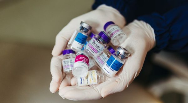 Medicininėmis pirštinėmis mūvinčios rankos laiko saują įvairių botulino buteliukų su skirtingomis etiketėmis, pexels.com nuotr.