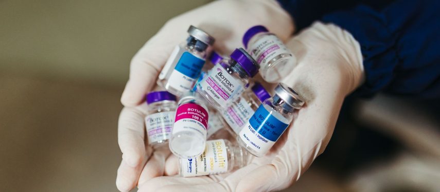 Medicininėmis pirštinėmis mūvinčios rankos laiko saują įvairių botulino buteliukų su skirtingomis etiketėmis, pexels.com nuotr.