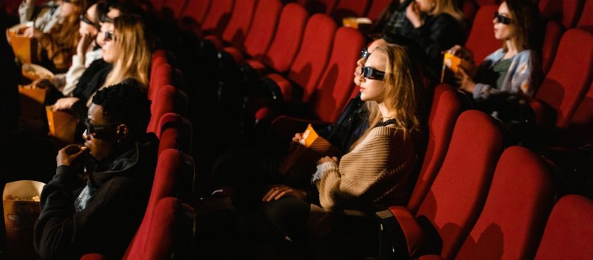 Kino salėje sėdi žiūrovai raudonose kėdėse, dėvi 3D akinius ir valgo spragėsius, pexels.com nuotr.